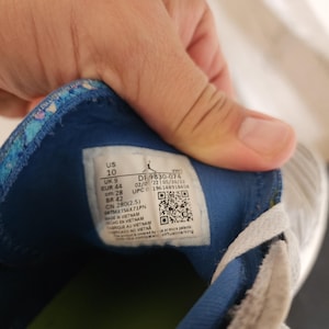 Puede incluir: Primer plano del interior de una zapatilla deportiva gris y blanca. La zapatilla tiene un forro azul y estampado. Una etiqueta muestra la talla EUR 44 y cm 28. La zapatilla est&aacute; fabricada en Vietnam.
