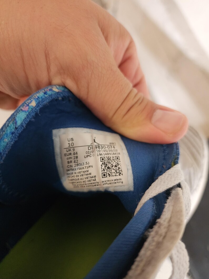 Puede incluir: Primer plano del interior de una zapatilla deportiva gris y blanca. La zapatilla tiene un forro azul y estampado. Una etiqueta muestra la talla EUR 44 y cm 28. La zapatilla est&aacute; fabricada en Vietnam.