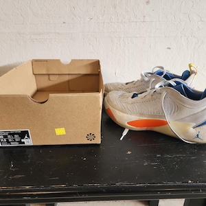 Puede incluir: Un par de zapatillas deportivas beige y azules con detalles en naranja, junto a una caja de zapatos de cart&oacute;n marr&oacute;n. Las zapatillas tienen cordones blancos y un logotipo de Jumpman visible. La caja tiene una etiqueta con informaci&oacute;n de tallas.
