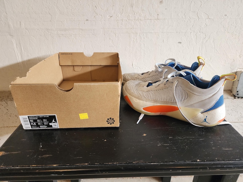 Puede incluir: Un par de zapatillas deportivas beige y azules con detalles en naranja, junto a una caja de zapatos de cart&oacute;n marr&oacute;n. Las zapatillas tienen cordones blancos y un logotipo de Jumpman visible. La caja tiene una etiqueta con informaci&oacute;n de tallas.