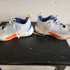 Puede incluir: Un par de zapatillas deportivas blancas y grises con detalles en naranja y azul. Las zapatillas tienen cordones blancos, un interior azul y una presilla amarilla en la parte trasera. Un peque&ntilde;o logotipo azul es visible en el lateral de cada zapatilla.