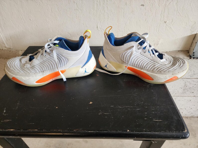 Puede incluir: Un par de zapatillas deportivas blancas y grises con detalles en naranja y azul. Las zapatillas tienen cordones blancos, un interior azul y una presilla amarilla en la parte trasera. Un peque&ntilde;o logotipo azul es visible en el lateral de cada zapatilla.