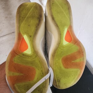 Puede incluir: Par de zapatillas deportivas, suelas hacia arriba, que muestran un patr&oacute;n de amarillo, naranja y blanco. Las suelas est&aacute;n sucias, con un dise&ntilde;o texturizado. Cordones blancos visibles.