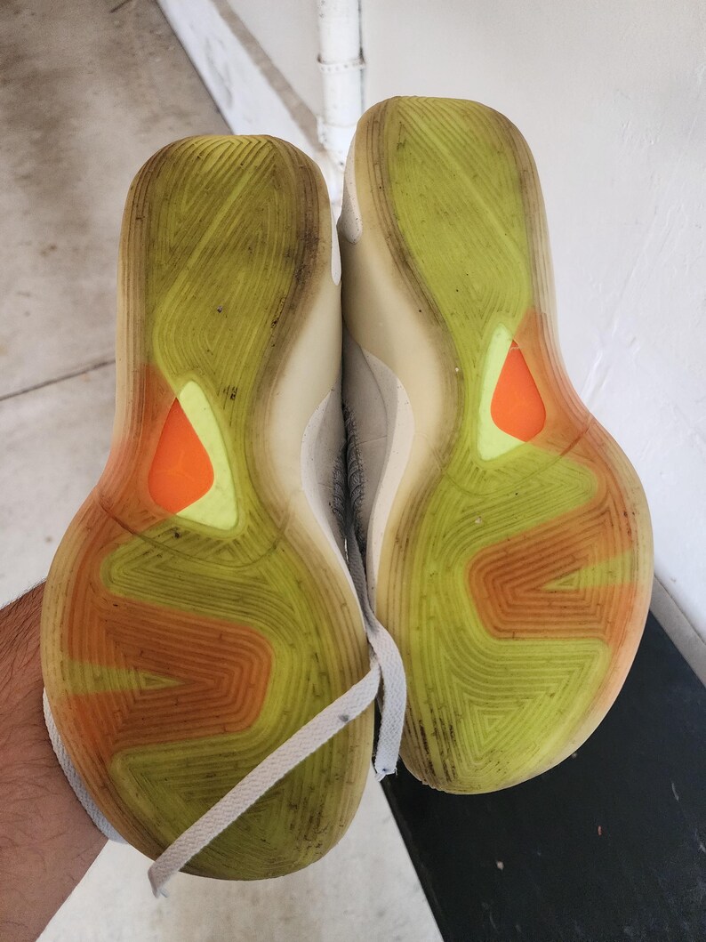 Puede incluir: Par de zapatillas deportivas, suelas hacia arriba, que muestran un patr&oacute;n de amarillo, naranja y blanco. Las suelas est&aacute;n sucias, con un dise&ntilde;o texturizado. Cordones blancos visibles.