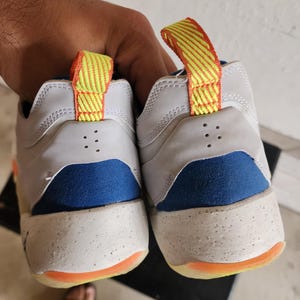 Puede incluir: Par de zapatillas blancas y azules con detalles en naranja y amarillo. Las zapatillas tienen una entresuela moteada y una leng&uuml;eta en el tal&oacute;n. Las suelas son naranjas.