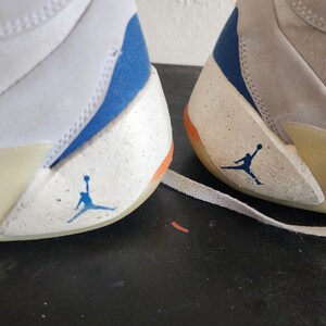 Puede incluir: Primer plano de dos zapatillas blancas y grises con detalles en azul y naranja. Las zapatillas presentan el ic&oacute;nico logotipo de Jumpman en azul en el lateral. El dise&ntilde;o incluye un patr&oacute;n moteado en las secciones blancas.