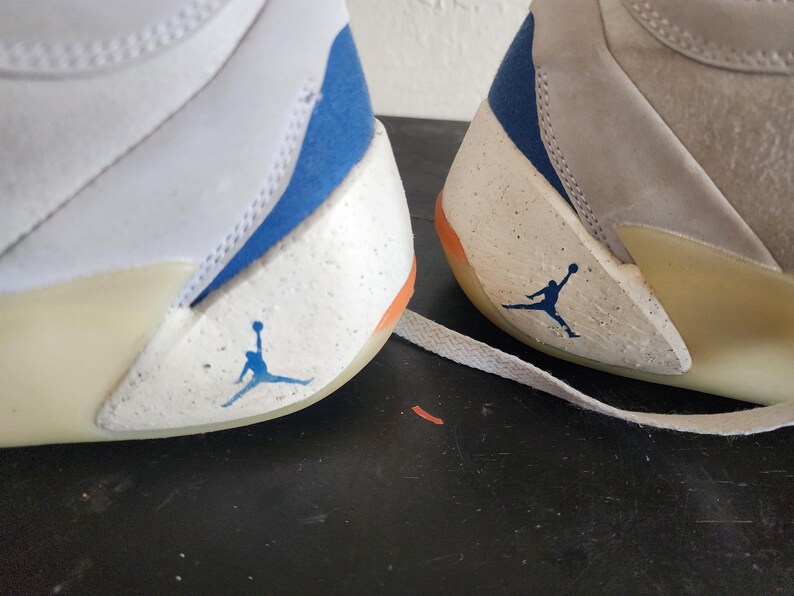 Puede incluir: Primer plano de dos zapatillas blancas y grises con detalles en azul y naranja. Las zapatillas presentan el ic&oacute;nico logotipo de Jumpman en azul en el lateral. El dise&ntilde;o incluye un patr&oacute;n moteado en las secciones blancas.