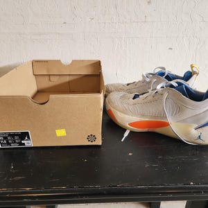 Puede incluir: Un par de zapatillas deportivas beige y azules con detalles en naranja, junto a una caja de zapatos de cart&oacute;n marr&oacute;n. Las zapatillas tienen cordones blancos y un logotipo azul de Jumpman. La caja tiene una etiqueta con informaci&oacute;n sobre la talla.