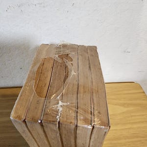 May include: A stack of wooden coasters, wrapped in clear plastic. The coasters are a light brown color and have a geometric shape. The packaging is slightly torn in places. The coasters are on a wooden surface.