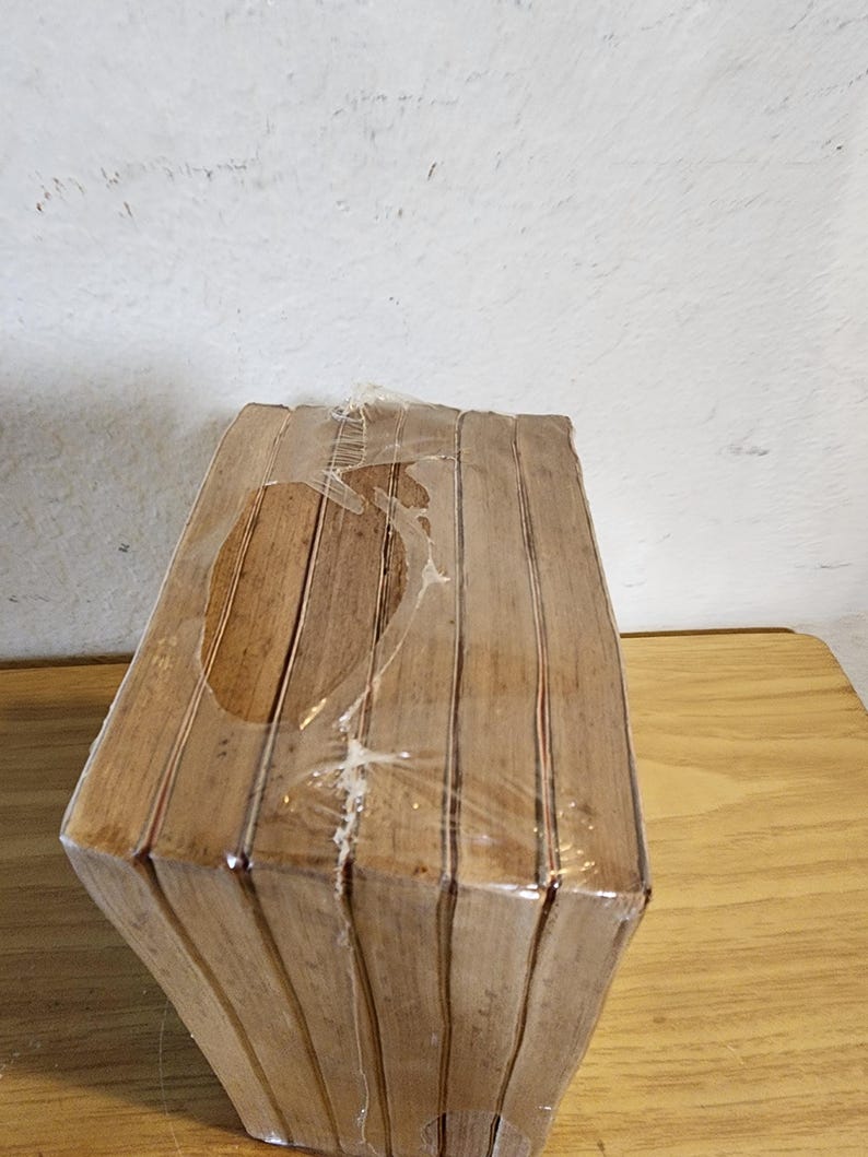 May include: A stack of wooden coasters, wrapped in clear plastic. The coasters are a light brown color and have a geometric shape. The packaging is slightly torn in places. The coasters are on a wooden surface.