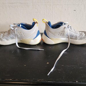 Puede incluir: Par de zapatillas deportivas grises con cordones blancos. Los zapatos tienen detalles en azul y amarillo, y una suela de color claro. Los zapatos est&aacute;n sobre una superficie oscura.