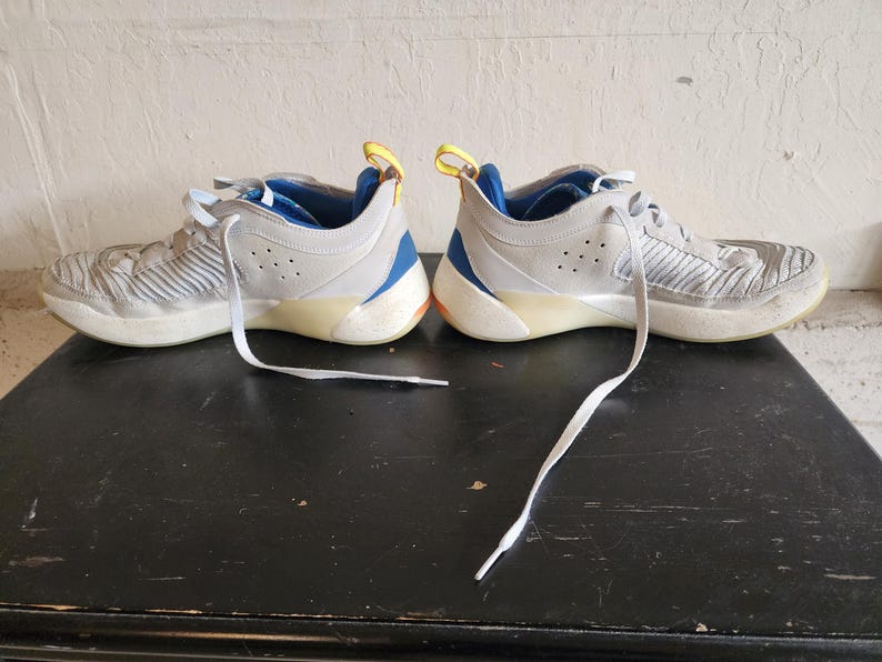 Puede incluir: Par de zapatillas deportivas grises con cordones blancos. Los zapatos tienen detalles en azul y amarillo, y una suela de color claro. Los zapatos est&aacute;n sobre una superficie oscura.