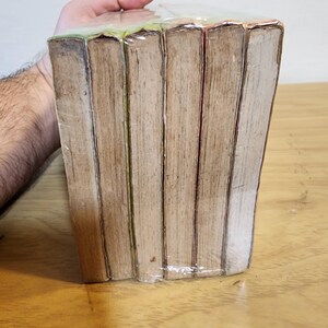 May include: A stack of books wrapped in clear plastic. The books have aged, light brown pages and spines. The edges of the pages are visible, and the books are held together by the plastic wrap. The books are on a wooden surface.
