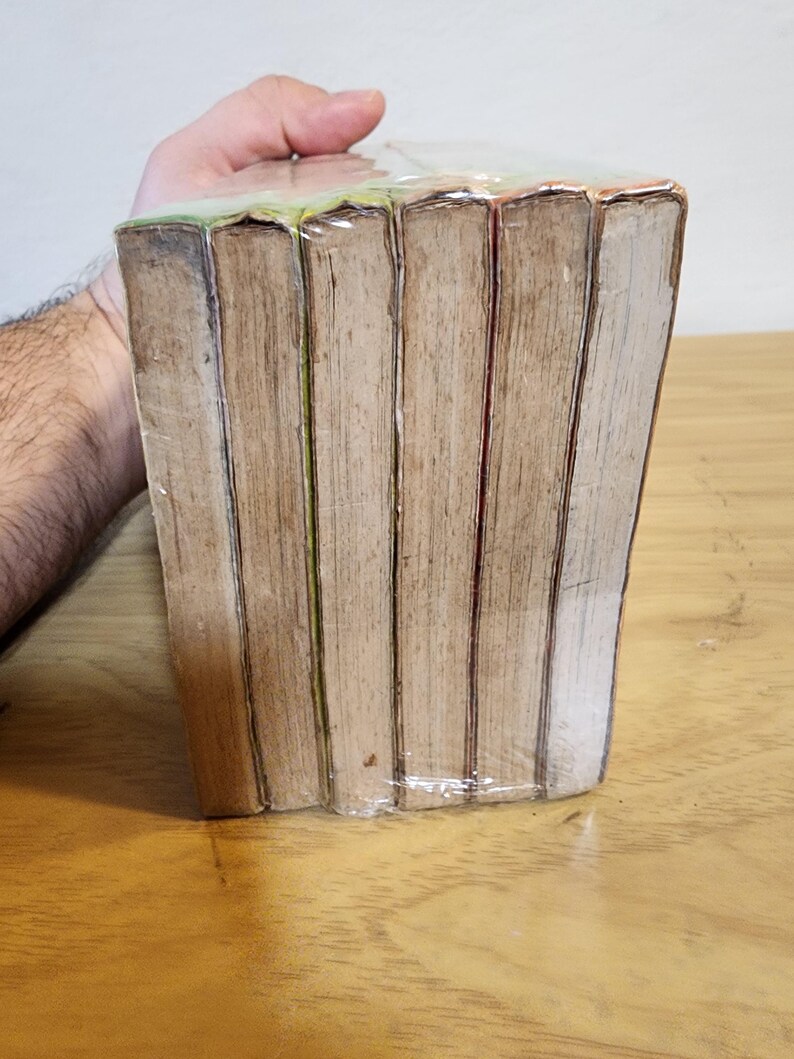 May include: A stack of books wrapped in clear plastic. The books have aged, light brown pages and spines. The edges of the pages are visible, and the books are held together by the plastic wrap. The books are on a wooden surface.