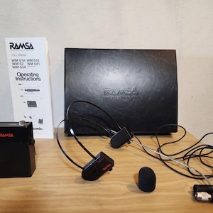 Op de afbeelding: Een zwart RAMSA draadloos microfoonsysteem met een headset, microfoon en ontvanger. De set bevat een handleiding en een zwarte draagtas. Het RAMSA-logo is zichtbaar op de componenten.