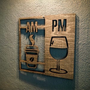 Könnte beinhalten: Wandkunstschild aus Holz mit den ausgeschnittenen Buchstaben "AM" und "PM". Die "AM"-Seite zeigt eine Kaffeetasse, die "PM"-Seite ein Weinglas. Das Schild ist aus hellbraunem Holz gefertigt und an einer strukturierten blauen Wand befestigt.