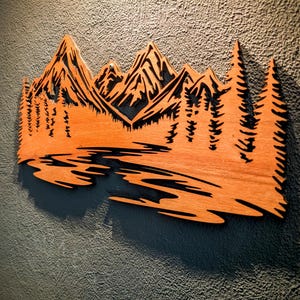Può includere: Opera d'arte da parete in legno raffigurante un paesaggio montano con alberi e un fiume. L'opera è tagliata dal legno e presenta una finitura naturale con venature del legno e dettagli neri. Montata su una parete grigia strutturata.