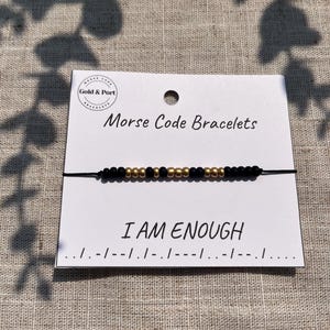 May include: A black string bracelet with gold and black beads spelling "I AM ENOUGH" in Morse code. The bracelet is displayed on a white card with the words "Morse Code Bracelets" printed on it. The brand name "Gold & Port" is also visible.