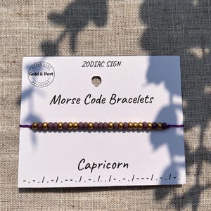 Peut inclure: Un bracelet signe du zodiaque Capricorne en code Morse, composé de perles violettes et dorées sur un cordon violet. Le bracelet est présenté sur une carte blanche avec le texte "ZODIAC SIGN", "Morse Code Bracelets" et "Capricorn".