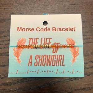 May include: A Morse code bracelet with gold and orange beads on a teal cord. The card behind the bracelet reads "Morse Code Bracelet" and "THE LIFE OF A SHOWGIRL" with Morse code symbols.