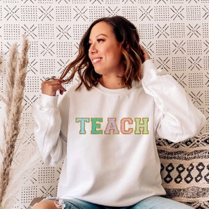 Op de afbeelding: Witte sweatshirt met het woord "TEACH" in kleurrijke, glinsterende letters. De letters zijn omlijnd met goud. De sweatshirt heeft een ronde hals.