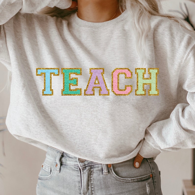 Op de afbeelding: Een lichtgrijze sweatshirt met het woord "TEACH" in kleurrijke, glinsterende letters. De letters zijn omlijnd in goud en gevuld met pastelkleuren: blauw, groen, paars en roze. De sweatshirt heeft een casual stijl.