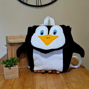 Peut inclure: Un sac à dos en forme de pingouin noir, blanc et jaune, avec des yeux bleus et un bec jaune. Il possède une poignée et des bretelles blanches, ainsi que des pieds bruns. Le sac à dos est posé sur une surface en bois.