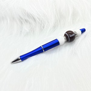 May include: A blue pen with a white and brown football-shaped bead. The bead is decorated with rhinestones.