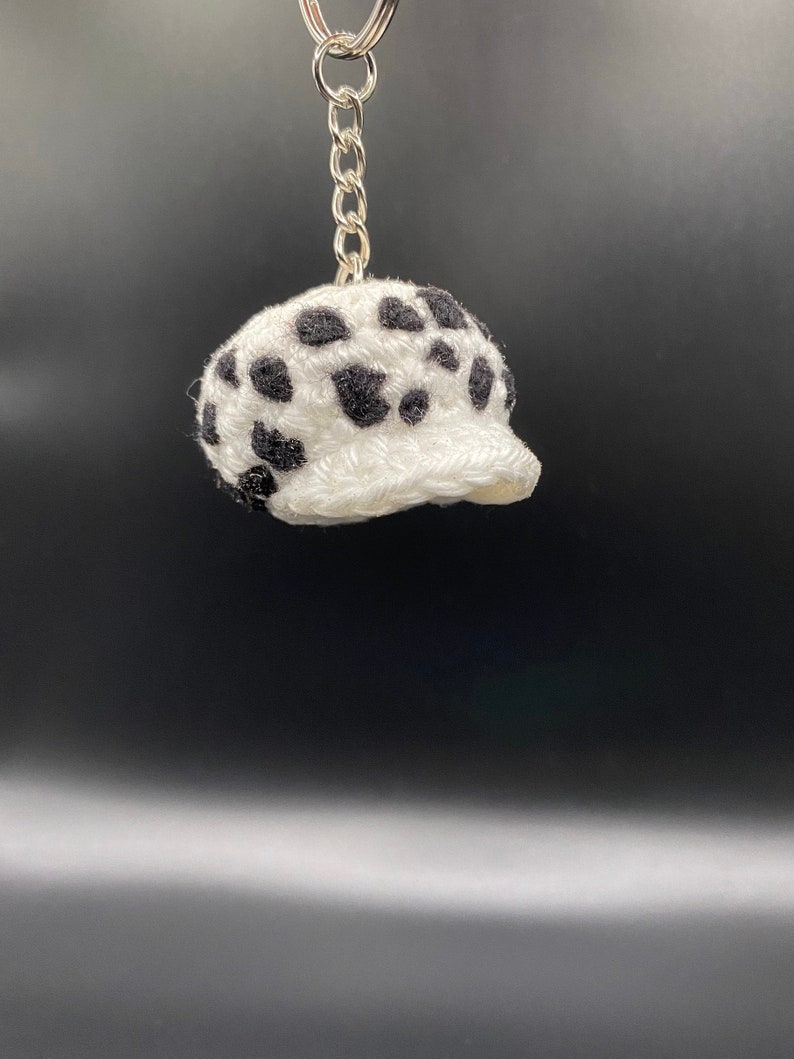 May include: White crocheted hat with black spots, shaped like a baseball cap, attached to a silver keychain.