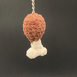May include: A brown and white crochet chicken leg keychain. The top portion is a textured brown, resembling fried chicken, while the bottom is a fluffy white, mimicking the bone. A silver chain and ring are attached for easy use.