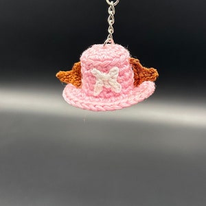 May include: A pink crocheted hat with brown wings and a white X on the front. The hat is attached to a silver key chain.