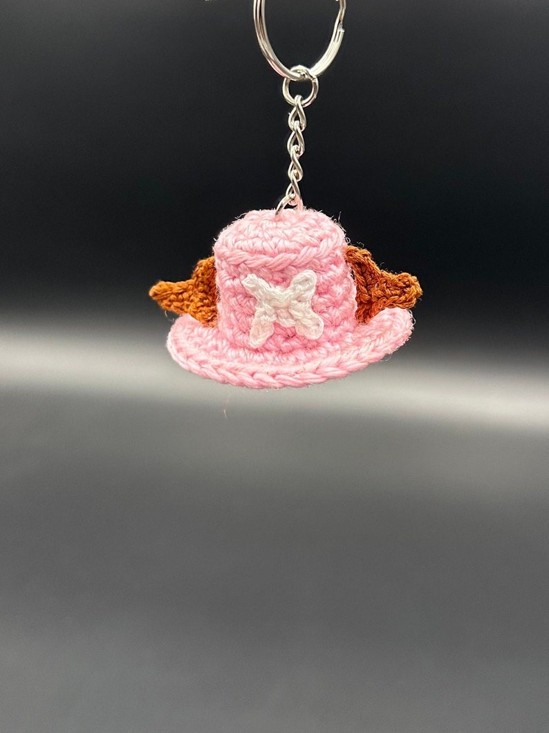 May include: A pink crocheted hat with brown wings and a white X on the front. The hat is attached to a silver key chain.