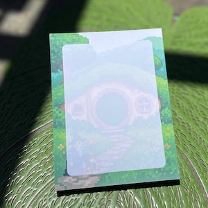 May include: A rectangular notepad featuring pixel art of a round-door dwelling nestled in a green landscape. The center of the paper has a light, semi-transparent rectangular area for writing, surrounded by a vibrant green border with pixelated trees.
