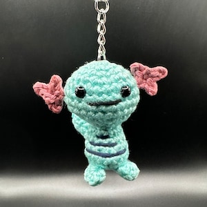 May include: A crocheted blue axolotl keychain with pink gills and black eyes. The keychain is attached to a silver key ring.