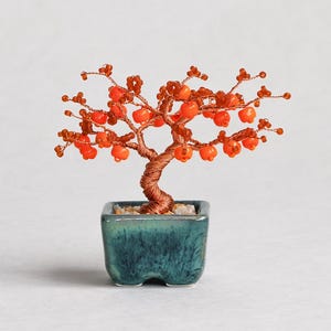 May include: A miniature wire bonsai tree sculpture featuring a twisted copper-colored trunk and branches adorned with small, bright orange beads. The tree is set in a small, square, teal-colored ceramic pot with a mottled, dark green glaze finish.
