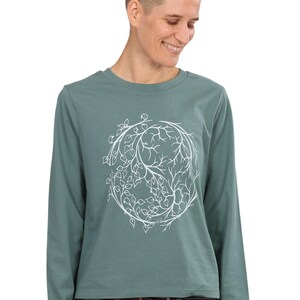 A person wearing a green long-sleeved shirt with a white tree design on it, smiling at the camera.