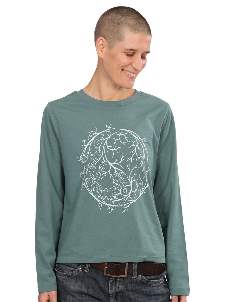A person wearing a green long-sleeved shirt with a white tree design on it, smiling at the camera.