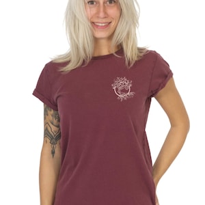May include: Burgundy crew neck t-shirt with a white tree of life graphic on the chest. The short-sleeved shirt has rolled cuffs and a curved hem. The model is wearing black pants.
