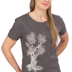 Könnte beinhalten: Graues T-Shirt mit einer detaillierten weißen Illustration eines Baumes und einem Vogel. Das Shirt hat einen Rundhalsausschnitt und kurze Ärmel. Das Model trägt eine schwarze Jeans.