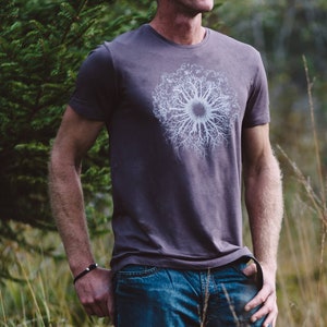 Könnte beinhalten: Ein Mann trägt ein dunkelgraues T-Shirt mit einem weißen Aufdruck eines Baumwurzelsystems. Er trägt eine blaue Jeans und ein schwarzes Armband am Handgelenk.