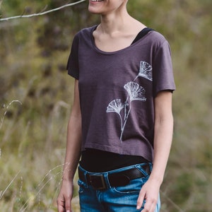 Può includere: Una donna indossa una maglietta corta color viola scuro con un design a foglia di ginkgo bianco. Indossa jeans blu e una cintura marrone.