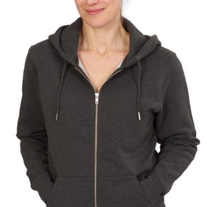 A woman wearing a gray hooded sweatshirt with her hands in her pockets, smiling at the camera.