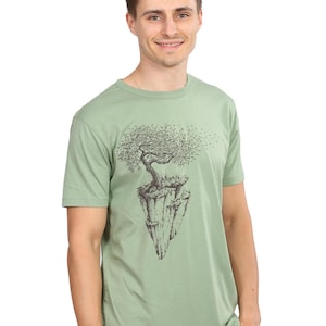 Könnte beinhalten: Salbeigrünes T-Shirt mit einer schwarz-weißen Illustration eines Baumes auf einem schwebenden Felsen. Das kurzärmelige T-Shirt ist aus weichem Material und für den lässigen Gebrauch geeignet. Das Design ist detailliert.