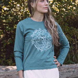 A woman with long blonde hair stands in front of a tree, wearing a teal long-sleeved shirt with a white tree design.