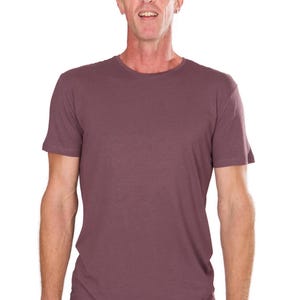 Könnte beinhalten: Ein kurzärmeliges T-Shirt mit Rundhalsausschnitt in einem gedämpften Malvenfarbton. Das Shirt hat ein schlichtes, klassisches Design und ist aus einem weichen, leichten Stoff gefertigt. Das Model trägt schwarze Shorts und ein Armband.