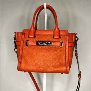 May include: A vibrant orange handbag with a pebbled texture, featuring two top handles and a detachable shoulder strap. The front is accented with a horizontal strap secured by a dark metallic turn-lock closure and matching side hardware.