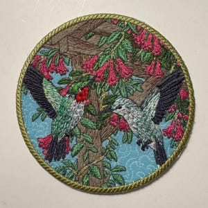 Könnte beinhalten: Gestickter Aufnäher mit zwei Kolibris mit grünen und weißen Federn, roten Kehlen und schwarzen Flügeln, vor dem Hintergrund rosa Blüten und einer Holzstruktur. Der Aufnäher hat einen hellblauen Hintergrund und einen grünen Rand.
