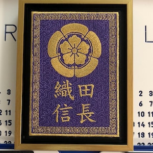 May include: A framed purple and gold embroidered wall hanging with the Japanese characters "織田信長" (Oda Nobunaga) in gold thread.