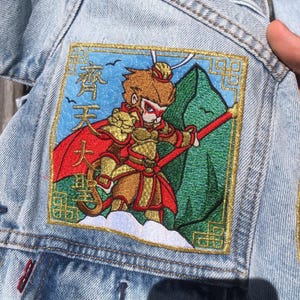 May include: Embroidered patch featuring a cartoon monkey warrior with a red cape and staff, set against a blue sky and green mountain backdrop. The patch is sewn onto a light blue denim jacket.