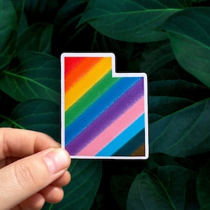 May include: A sticker in the shape of the state of Utah, featuring a rainbow gradient design. The sticker has a white border and is held against a backdrop of green leaves.